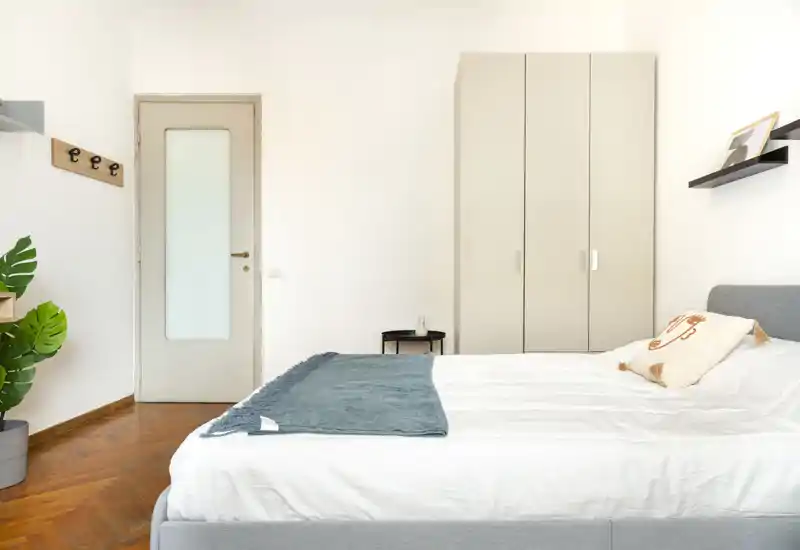 Camera da letto luminosa e moderna con un ampio letto con biancheria bianca, un plaid accogliente e un armadio a muro. Arredamento minimale e pavimento in legno scuro donano un’atmosfera invitante, perfetta per annunci di affitto.