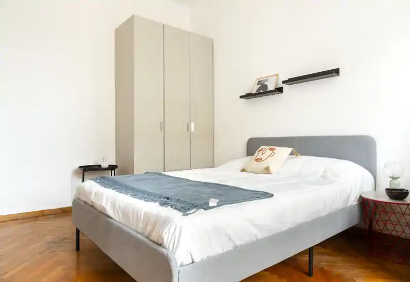 Camera da letto luminosa e minimalista con un letto matrimoniale ben rifatto, testiera grigio chiaro, pavimento in parquet e un armadio alto; toni neutri e arredi semplici per un’atmosfera riposante adatta agli annunci.