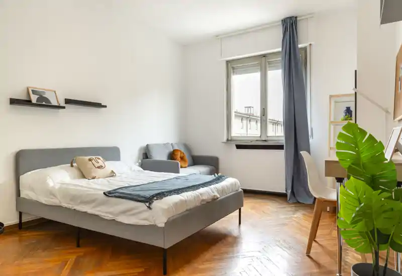 Camera da letto luminosa e moderna con letto imbottito grigio, pavimento in parquet a spina di pesce, una poltroncina e una scrivania vicino alla finestra—ben arredata e illuminata naturalmente, perfetta per annunci.