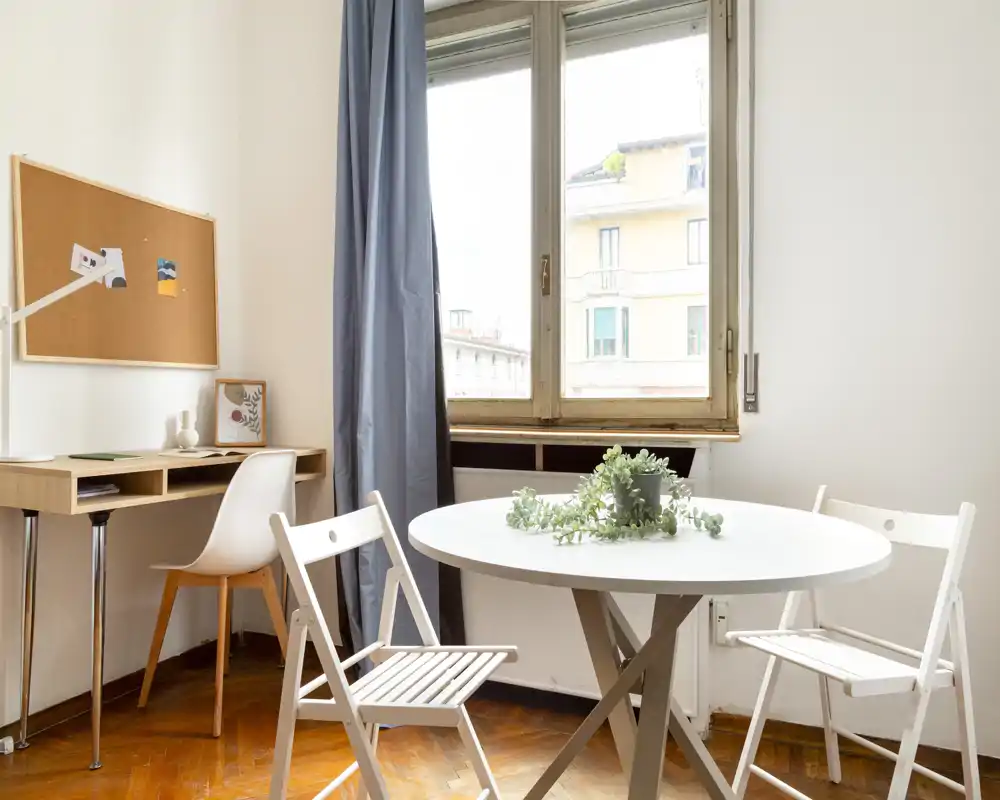 Zona pranzo luminosa e minimal con tavolo rotondo bianco, sedie pieghevoli e una pianta decorativa vicino a una grande finestra.