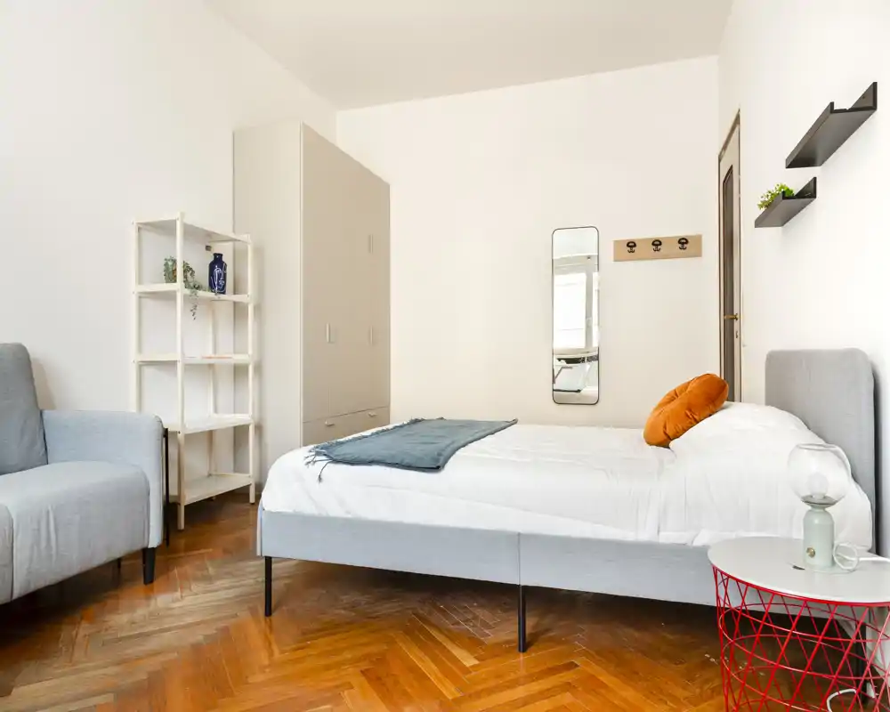 Camera da letto luminosa e moderna con letto matrimoniale confortevole, tessili morbidi e pavimento in parquet caldo. Arredamento essenziale con guardaroba, scaffale e piccolo angolo salotto.