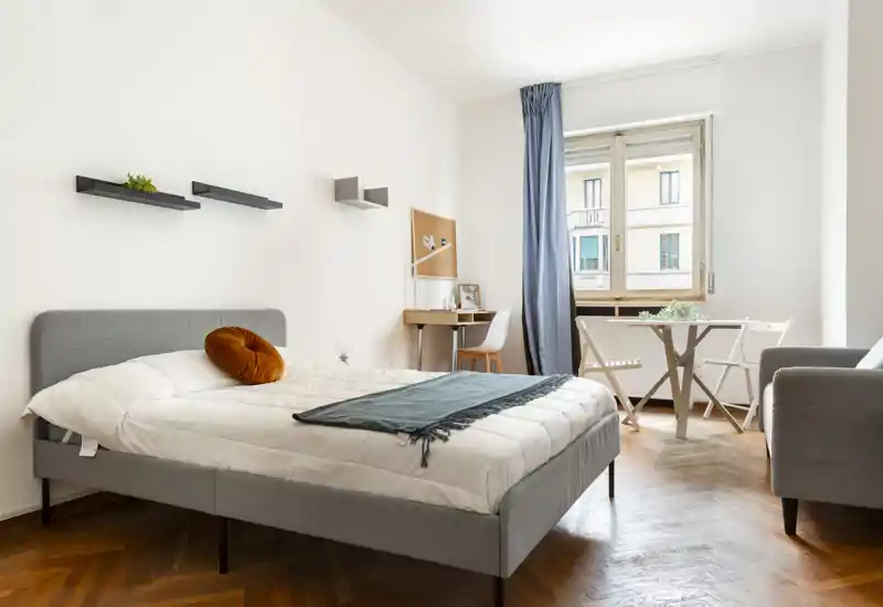 Camera da letto luminosa e moderna con letto imbottito grigio, biancheria morbida, cuscino decorativo rotondo e una piccola scrivania vicino alla finestra. Il pavimento in legno e la luce naturale creano un’atmosfera calda e accogliente, perfetta per annunci.