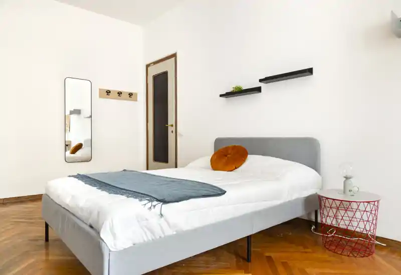 Camera da letto luminosa e moderna con letto imbottito grigio completamente visibile, pavimento in parquet caldo, cuscino arancione rotondo decorativo e un plaid blu. Mensole minimal e uno specchio a figura intera completano l'allestimento elegante.