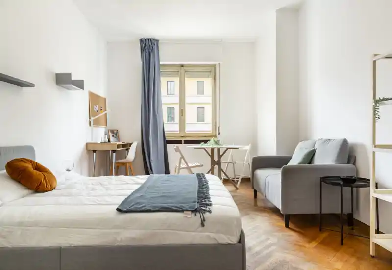 Camera da letto moderna e luminosa con ampissimo letto in primo piano, tessili morbidi e un cuscino decorativo; luce naturale dalla finestra e una piccola zona salotto con divanetto e tavolo rendono l'ambiente accogliente.