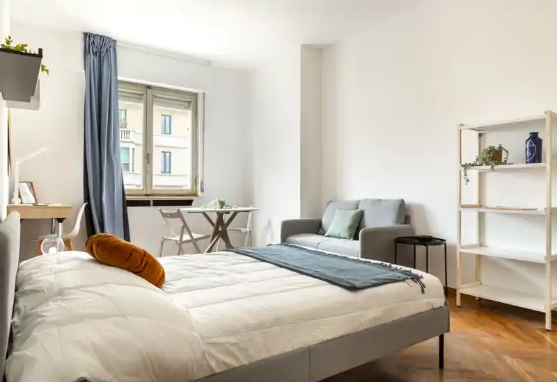 Dormitorio luminoso y bien decorado con una cama doble cómoda en primer plano, una zona de estar pequeña, escritorio junto a la ventana y suelos de madera—ideal para anuncios de piso.