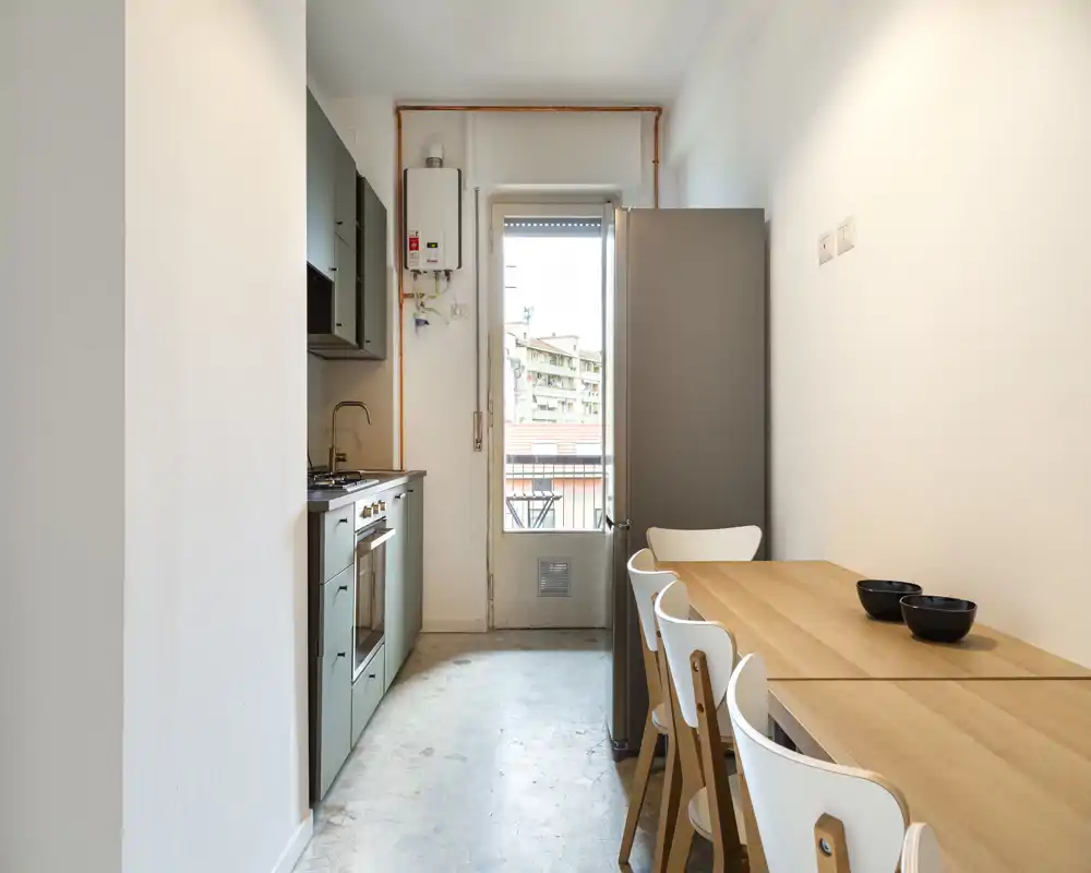 Cucina moderna compatta con forno integrato, piano cottura e un tavolo in legno con sedie accanto a una porta che si apre su un piccolo balcone — luminosa e ben arredata per la vita in città.
