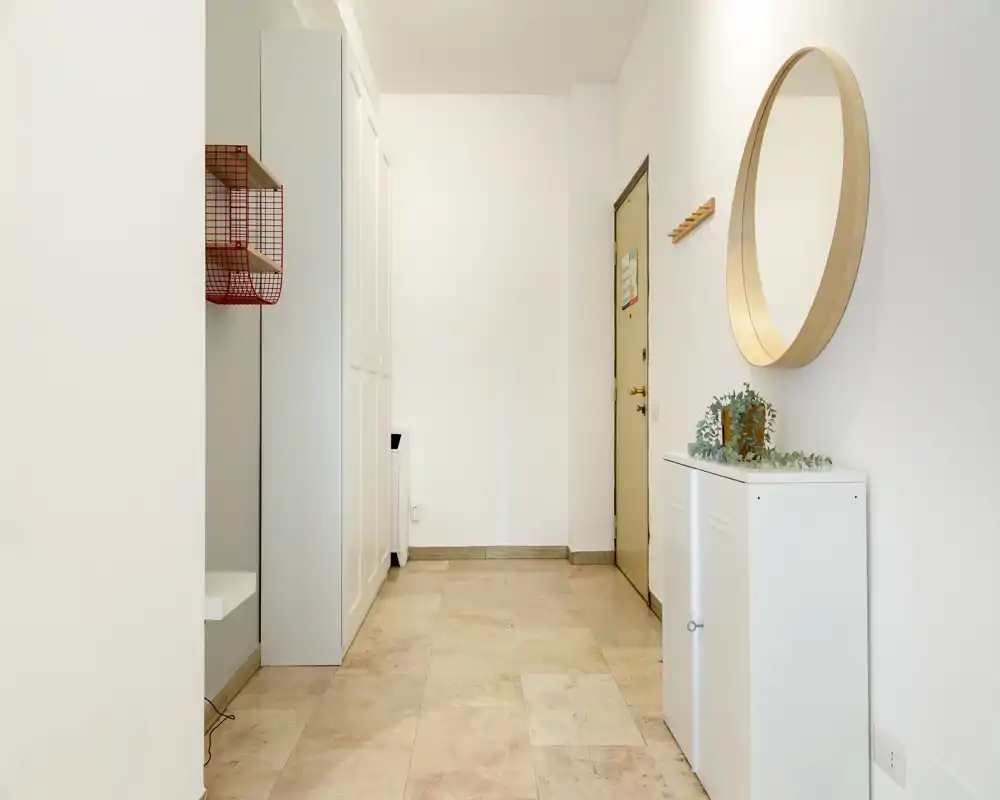 Ingresso luminoso e minimalista con specchio rotondo a parete, piccolo mobiletto e appendiabiti vicino alla porta — pavimento piastrellato e pareti neutre per un’atmosfera ordinata.