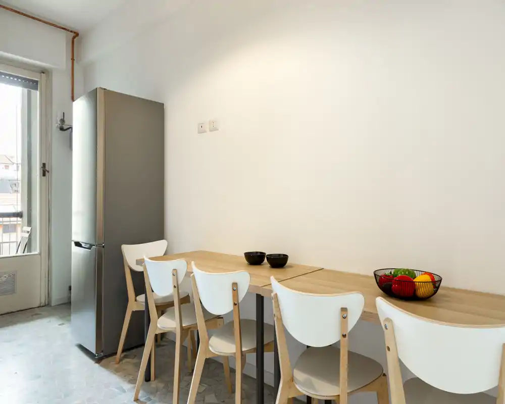 Angolo cucina luminoso e minimal con frigorifero alto in acciaio, piano in legno usato come zona pranzo e quattro sedie bianche — soluzione compatta e moderna per monolocali.