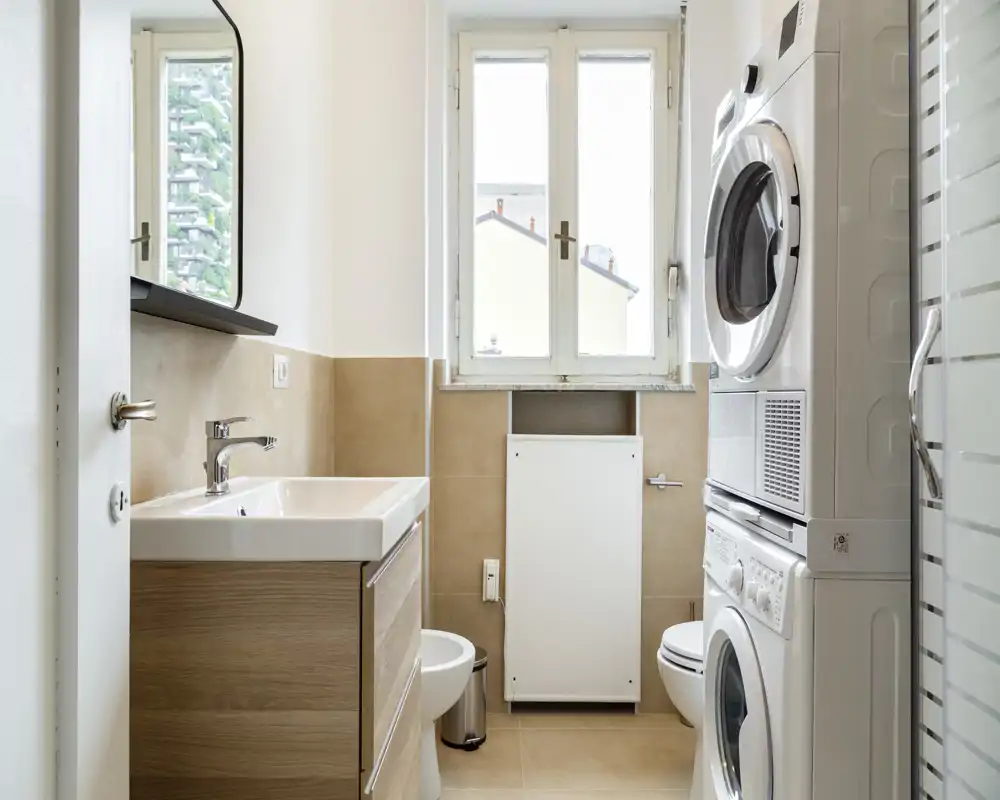 Bagno moderno e luminoso con lavabo, lavatrice e asciugatrice impilate, wc e una finestra che illumina l'ambiente.