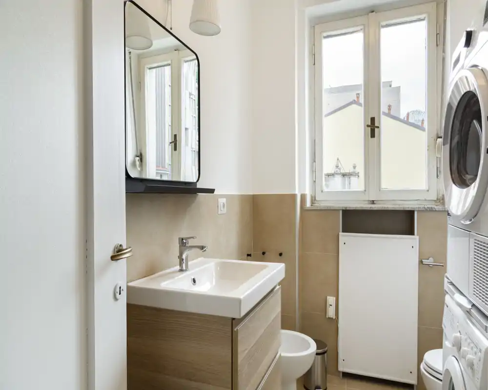 Bagno moderno e luminoso con specchio a parete, lavabo con mobile in legno, wc e colonna lavatrice-asciugatrice; piastrelle beige e una finestra ampia forniscono luce naturale.