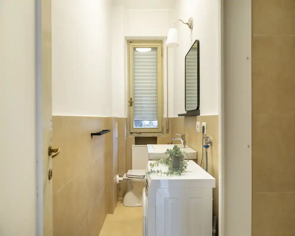 Bagno stretto e luminoso con WC, lavabo, specchio e lavatrice. Piastrelle beige neutre e una finestra che garantisce luce naturale gradevole.