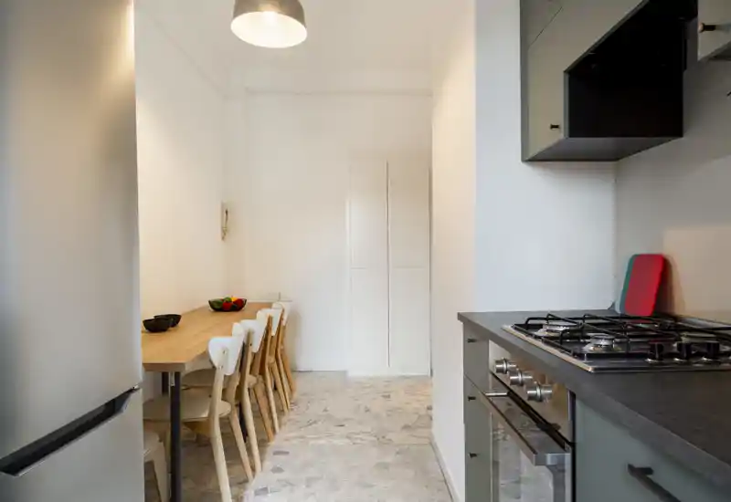 Cocina moderna con placa de gas y horno integrados en una encimera oscura, armarios a juego y una mesa de madera con sillas. Distribución luminosa y funcional, ideal para apartamentos urbanos compactos.