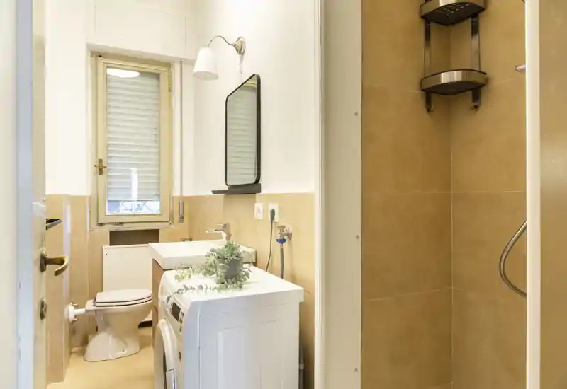 Baño compacto y luminoso con inodoro, lavabo sobre mueble, lavadora y plato de ducha alicatado. Baldosas beige neutras y una ventana pequeña que proporciona luz natural.