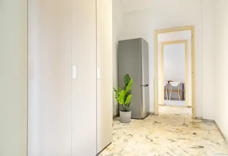 Entrada luminosa y minimalista con un armario alto a la izquierda, un frigorífico de acero y una planta en maceta junto a la puerta que conduce al comedor.