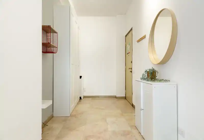 Entrada luminosa y minimalista con espejo redondo en la pared, pequeño mueble y percheros junto a la puerta — suelo de baldosas y paredes neutras que crean un recibidor ordenado.