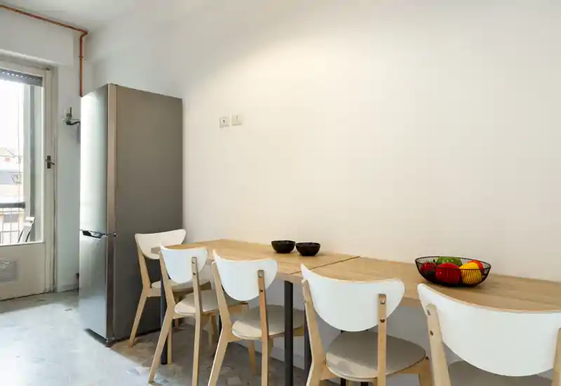 Rincón de cocina luminoso y minimalista con frigorífico alto de acero, encimera de madera usada como mesa y cuatro sillas blancas — configuración compacta y moderna ideal para pisos pequeños.