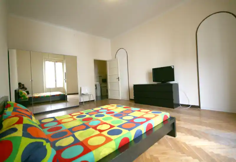 Amplio dormitorio con cama baja y edredón geométrico colorido, gran armario con puertas espejo, cómoda con televisor y suelo de parqué. Luminoso y con mobiliario mínimo, perfecto para alquiler moderno.
