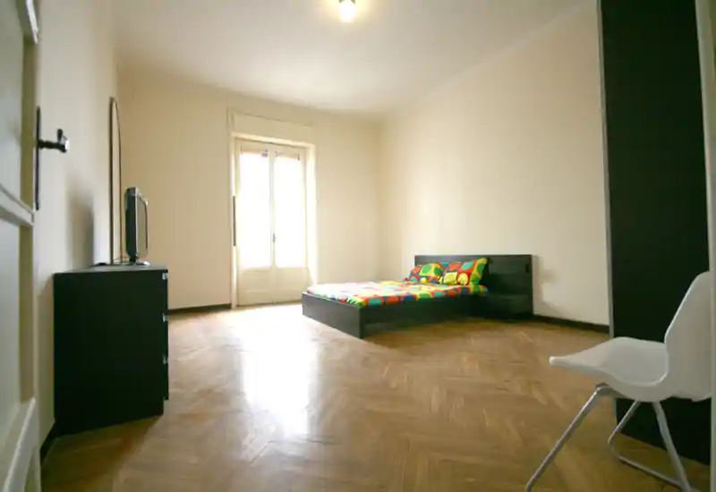 Amplio dormitorio con mobiliario sencillo y una cama baja con ropa de cama colorida, gran ventana/puerta que aporta luz natural y suelo de parquet.