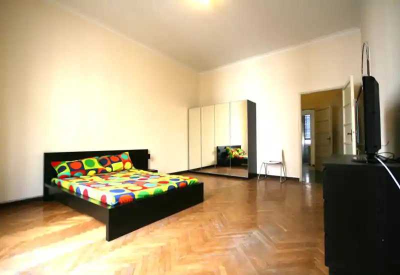Amplio dormitorio con cama plataforma negra y ropa de cama colorida, suelo de parquet y un gran armario con espejo que aporta sensación de amplitud y luz.
