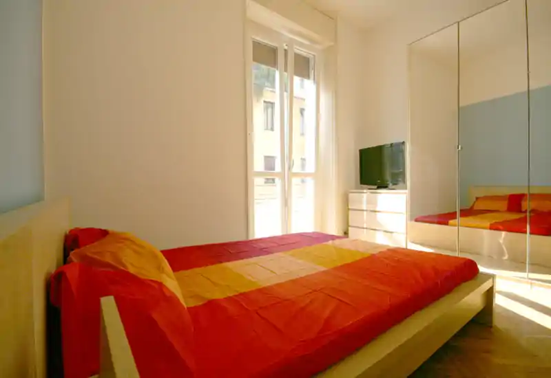 Chambre lumineuse avec lit double habillé de linge rouge et orange, grande fenêtre apportant de la lumière naturelle et placard à portes miroir qui agrandit l'espace.