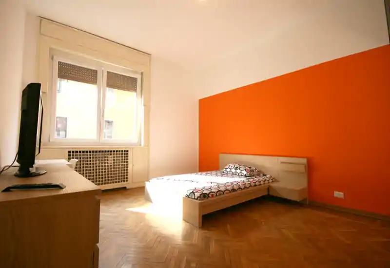 Dormitorio individual luminoso con suelo de parquet, pared de acento naranja y una cama baja moderna situada junto a una gran ventana que aporta abundante luz natural.