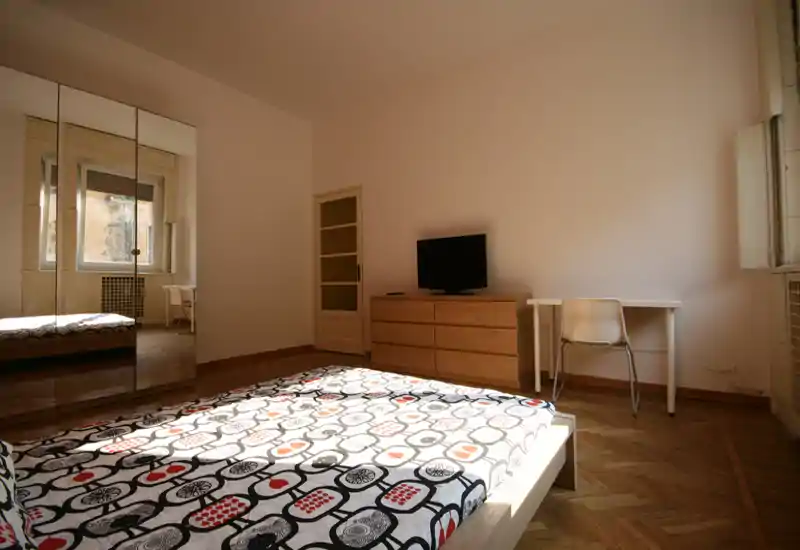 Amplio dormitorio con una cama grande en primer plano, armario con espejo, cómoda de madera con TV y un pequeño escritorio junto a la ventana — buena luz natural y suelo de parquet.