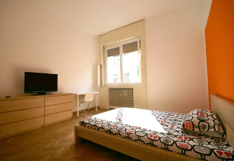 Dormitorio moderno y luminoso con cama doble, ropa de cama estampada y pared naranja como acento. Gran ventana que aporta abundante luz natural; cómoda y escritorio sencillos para almacenamiento y trabajo.