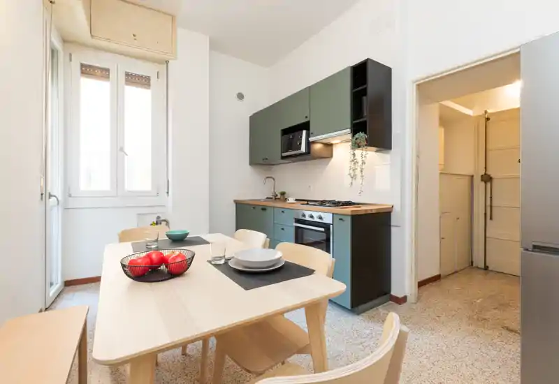 Cucina moderna e luminosa con mobili verdi, piano in legno e tavolo da pranzo apparecchiato con piatti e una ciotola di frutta — perfetta per annunci di affitto fotografici.