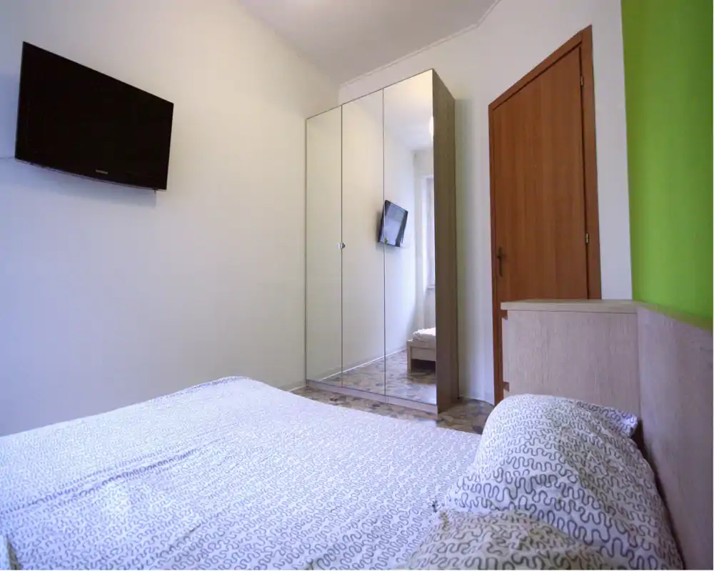 Camera luminosa e arredata in modo semplice con letto matrimoniale in primo piano, armadio con ante a specchio e TV a parete; tonalità neutre e luce naturale per un ambiente pulito e arioso.