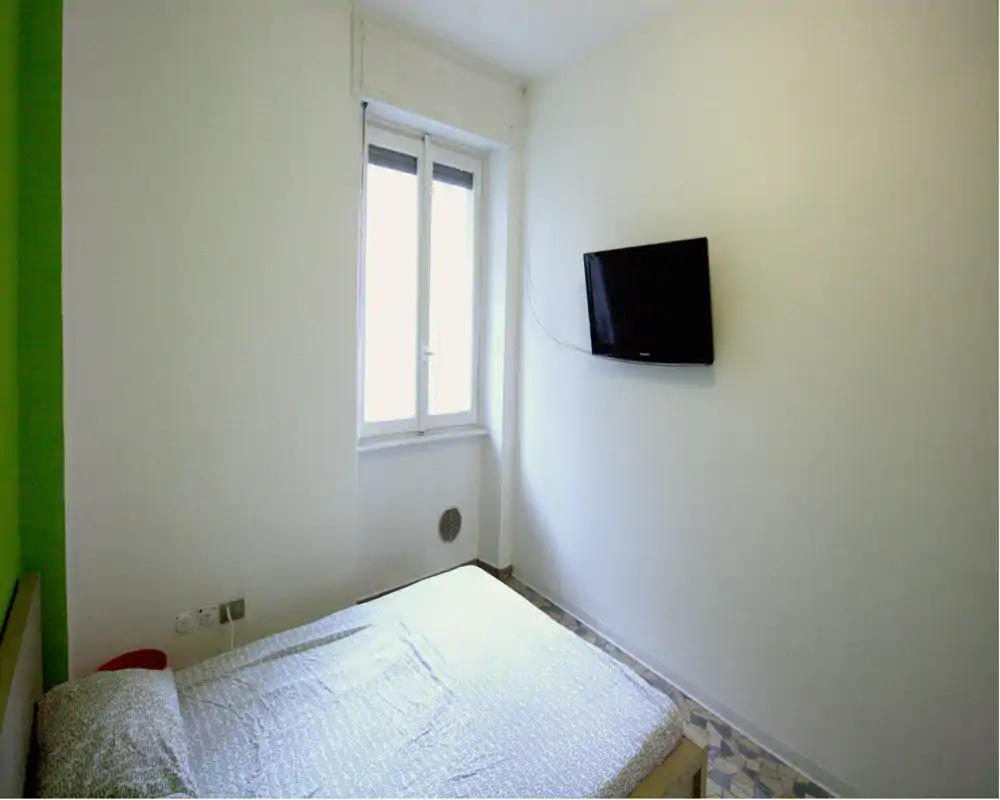 Camera da letto piccola con letto matrimoniale adiacente al muro, finestra che fornisce luce naturale e TV a parete. Ambiente semplice e luminoso, ideale per soggiorni brevi o affitti economici.