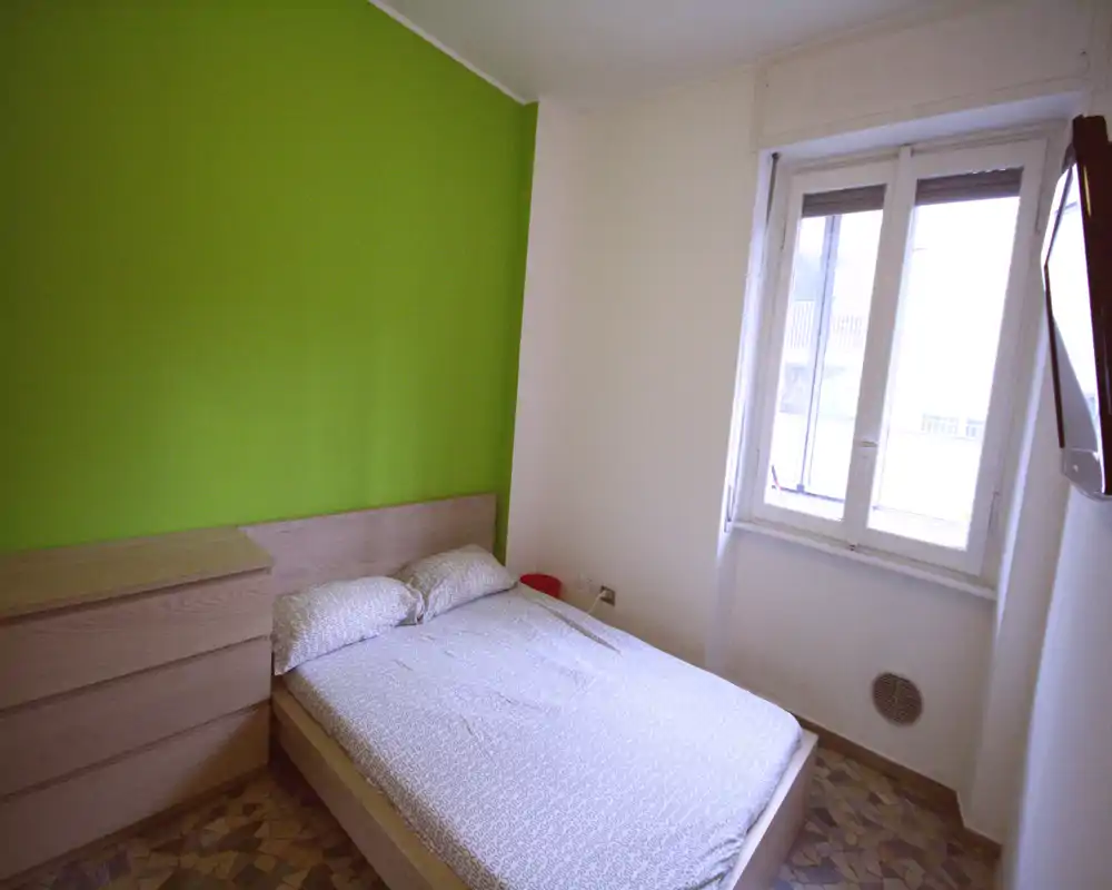 Camera matrimoniale luminosa con letto in legno chiaro, biancheria a fantasia e una parete verde brillante come punto focale. Ampia finestra che garantisce molta luce naturale e comò con cassetti per riporre gli effetti personali.