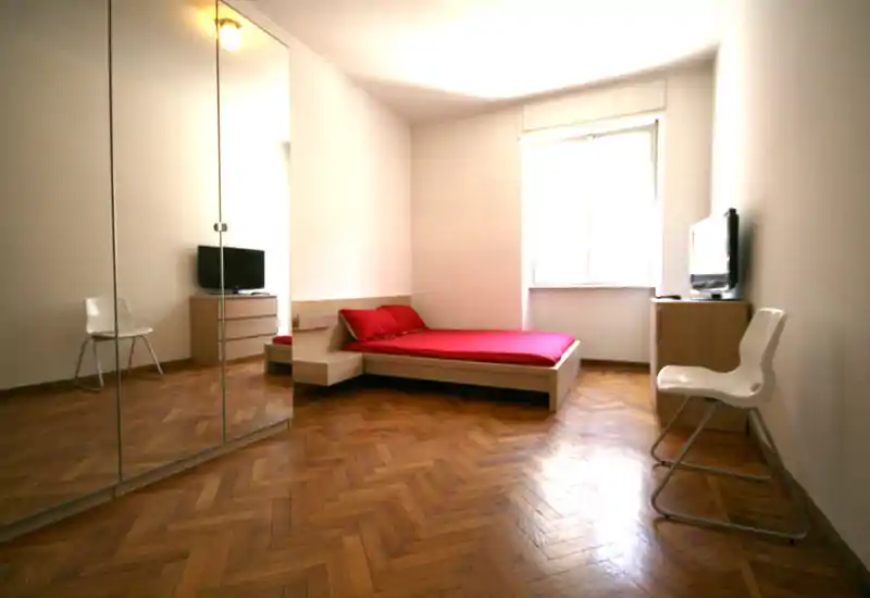 Bright, minimalist bedroom with a low platform bed dressed in red linens, parquet floors and a large mirrored wardrobe that reflects extra space.