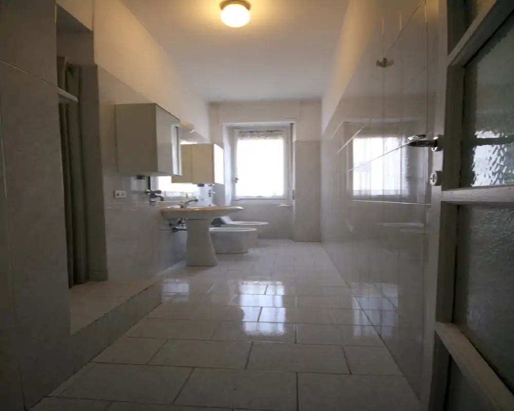 Bright, tiled bathroom with pedestal sink, bidet and toilet aligned toward a window; glossy floor and wall tiles create a clean, reflective look.