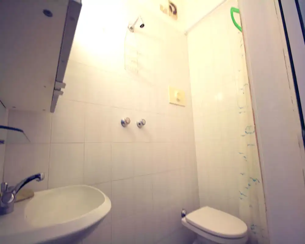 Compact bathroom with white tiled walls, a wall-mounted sink and a toilet; small shower area with a curtain is visible on the right.