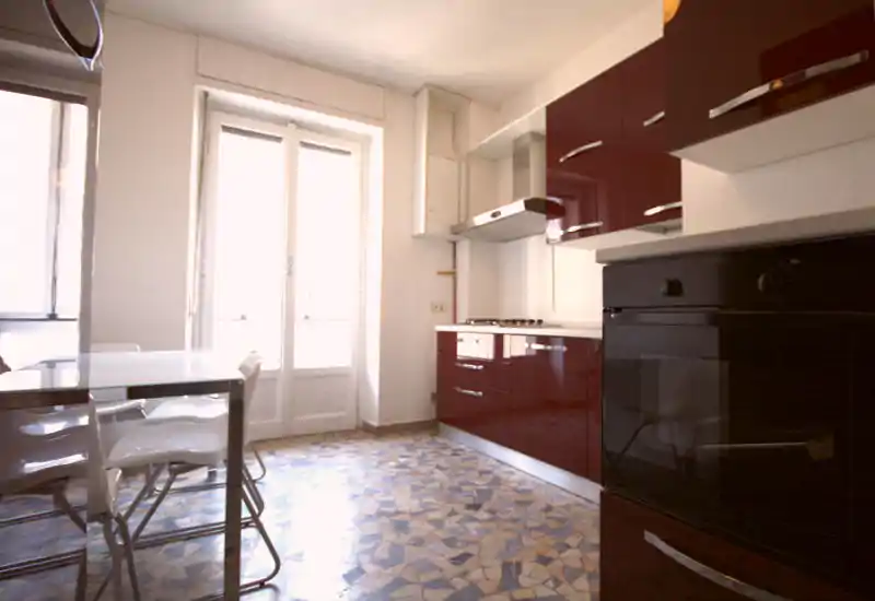 Bright modern kitchen with glossy burgundy cabinets, built-in oven and gas hob, plus a small dining table by a large window/door that floods the room with natural light.