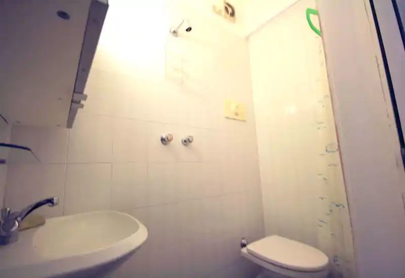 Compact bathroom with white tiled walls, a wall-mounted sink and a toilet; small shower area with a curtain is visible on the right.