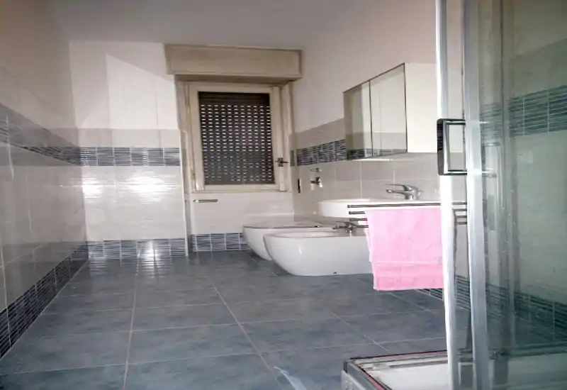 Bright, modern bathroom with blue floor tiles, wall-mounted sink, bidet and toilet, and a glass shower enclosure visible at right. Clean lines and neutral tones make it suitable for rental listings highlighting functionality.