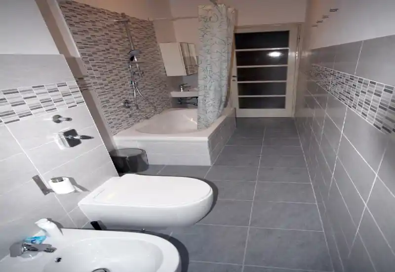 Narrow tiled bathroom with bathtub and shower, wall-mounted toilet and bidet. Modern grey tiles and mosaic accent strip create a cohesive, clean look.