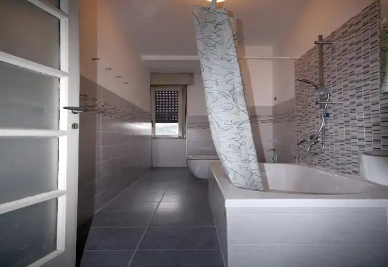 Bright narrow bathroom with bathtub and wall-mounted shower, modern grey tiles and patterned shower curtain; compact layout with a window at the end.