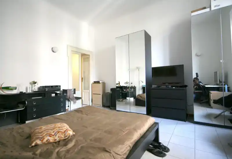 Camera da letto luminosa e moderna con un grande letto basso in primo piano, armadi con ante a specchio che ampliano lo spazio e mobili neri dal design minimale. Illuminazione naturale uniforme e linee pulite, ideale per annunci di affitto.