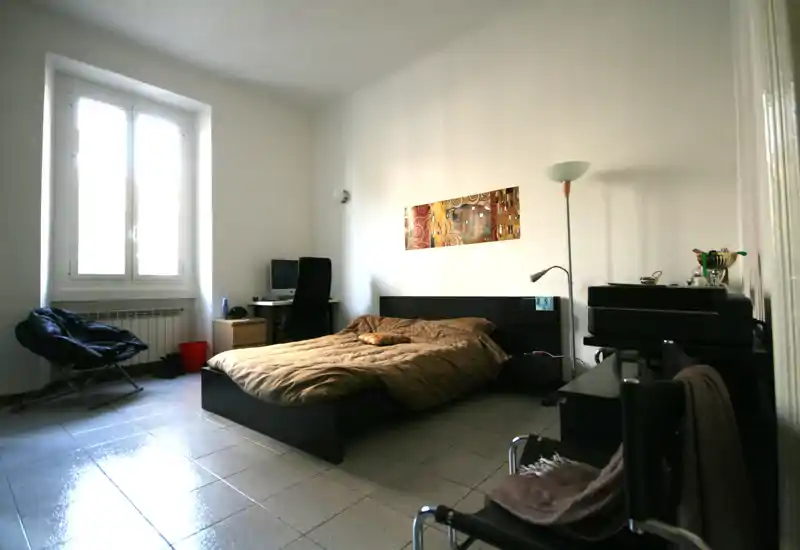 Camera da letto moderna e luminosa con letto basso scuro, piumone caldo e una grande finestra che fornisce luce naturale. Una postazione con scrivania e sedia è accanto al letto; arredi minimali e contemporanei mantengono la stanza ordinata.