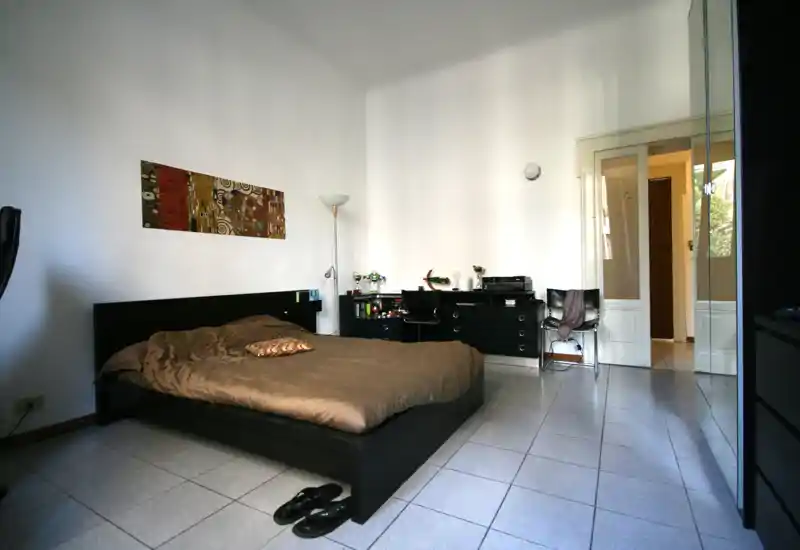 Ampia camera da letto con letto su piattaforma scuro, biancheria neutra e una scrivania/comò sulla parete opposta. Pareti bianche luminose e un'ampia porta creano un senso di spazio; un quadro decorativo e una lampada da terra aggiungono personalità.