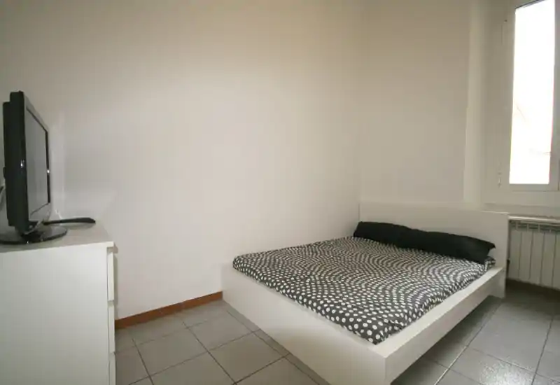 Chambre minimaliste avec lit blanc surélevé, couette à pois et fenêtre apportant de la lumière naturelle. Lignes épurées et mobilier simple, idéale pour les locations de courte durée.