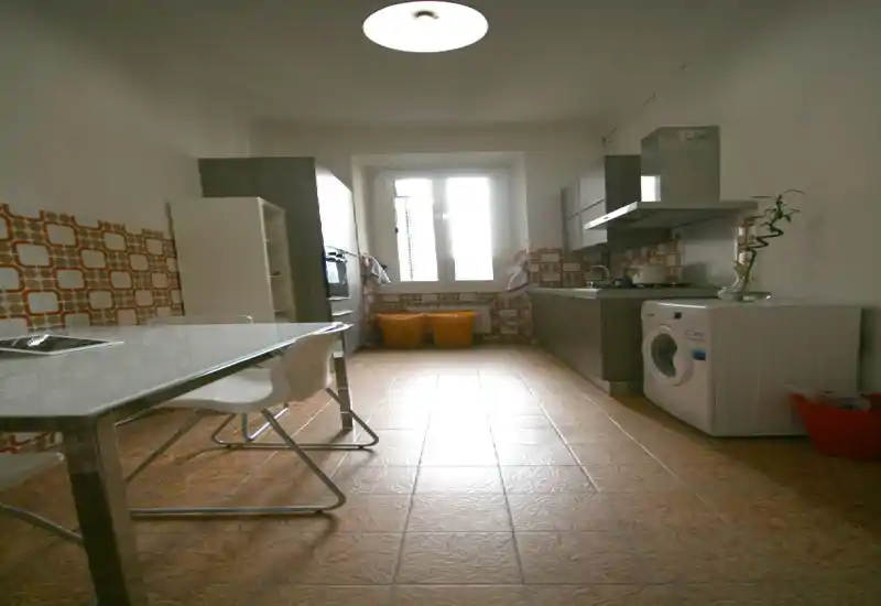 Cuisine en enfilade lumineuse avec carrelage décoratif mural et au sol, meubles bas modernes, hotte et lave-linge. Une table simple avec chaises blanches et une grande fenêtre apportent la lumière naturelle.