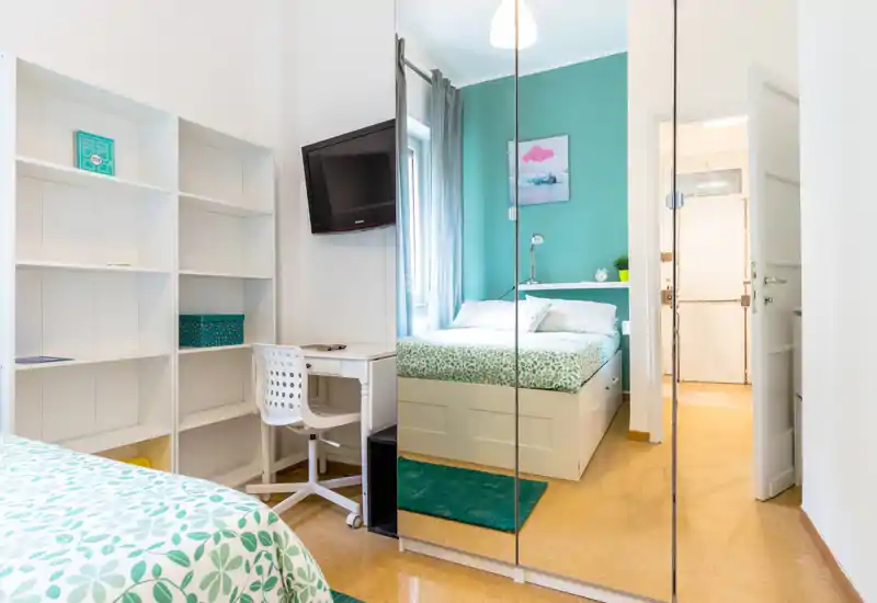 Camera da letto luminosa e ben curata con letto matrimoniale, armadio con ante a specchio e una scrivania vicino alla finestra. La parete verde e i tessuti coordinati donano un'atmosfera fresca e accogliente.