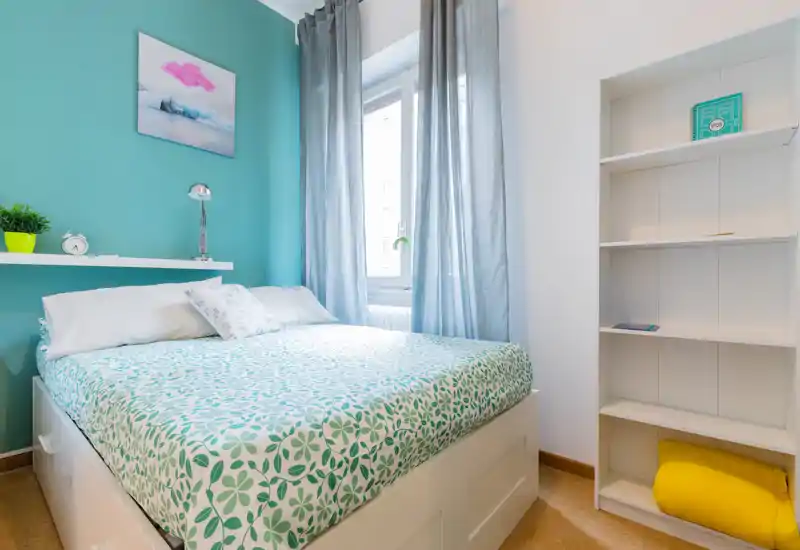 Camera da letto luminosa e moderna con letto matrimoniale decorato, morbidi cuscini e parete accentata turchese. La luce naturale dalla finestra e le mensole ordinate creano un’atmosfera fresca e accogliente, perfetta per annunci.