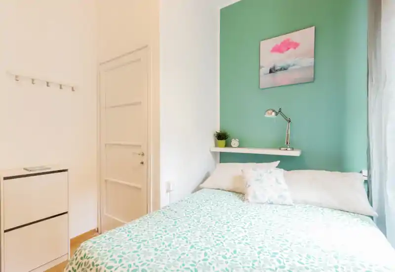 Camera da letto singola luminosa e accogliente con copriletto a motivo, cuscini decorativi, mensola con lampada e pianta e parete accentata verde menta.