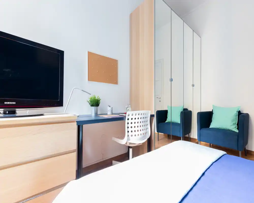 Camera da letto moderna e luminosa con letto ben visibile in primo piano, scrivania e TV a sinistra e un armadio con ante specchiate affiancato da una poltrona, layout funzionale.