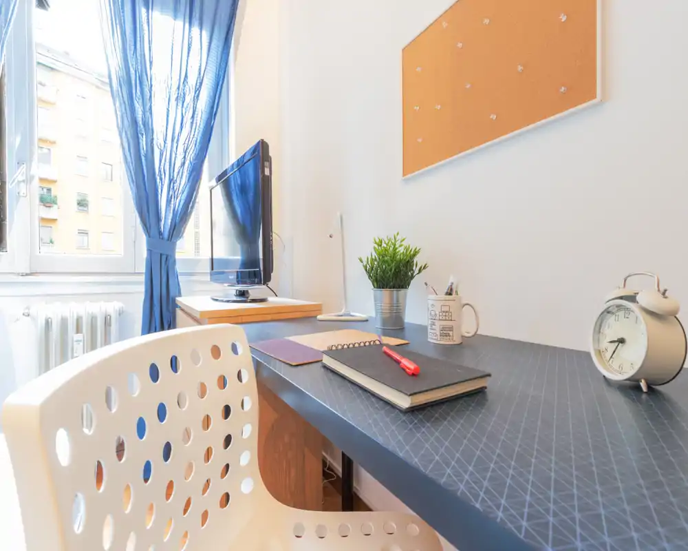 Spazioso angolo studio in camera da letto, luminoso e ordinato, con scrivania vicino alla finestra, pianta decorativa, bacheca e TV — ideale per studiare o lavorare da casa.