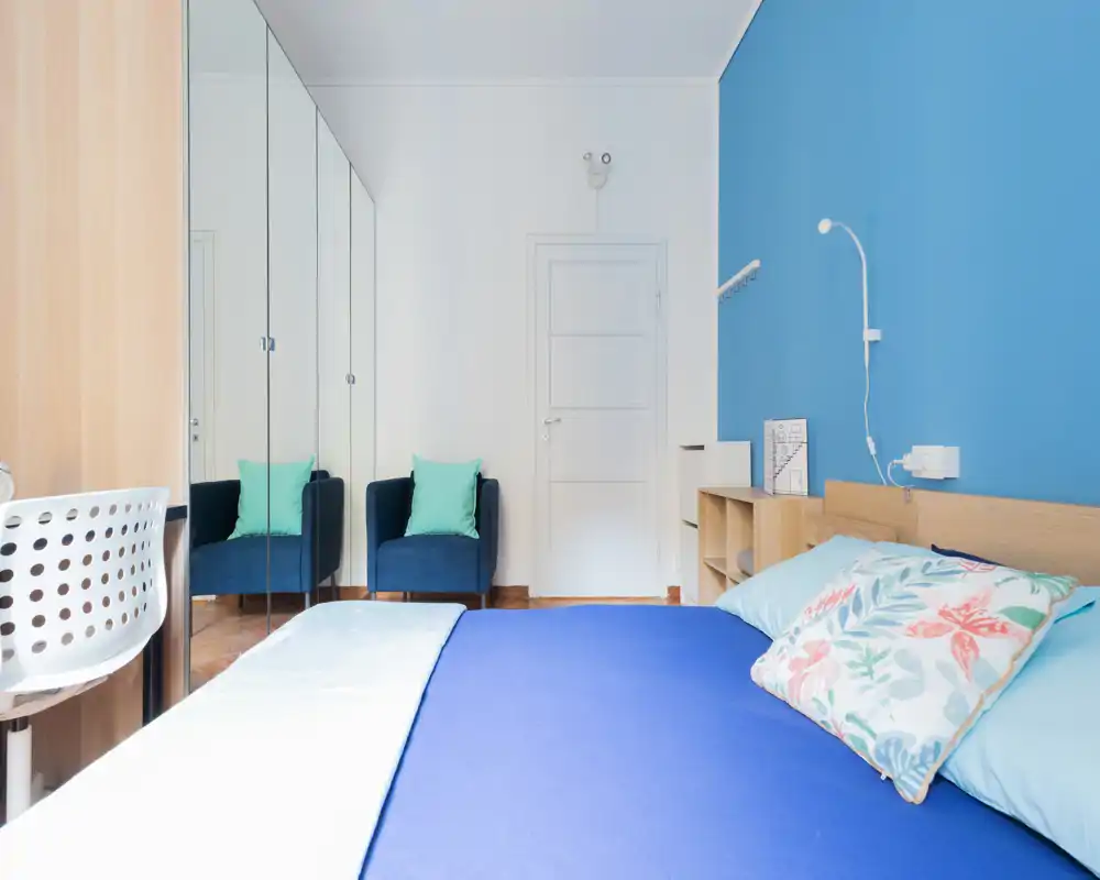 Camera da letto moderna e luminosa con un ampio letto blu in primo piano, cuscini decorativi, mensole accanto al letto e un armadio con specchi che aggiunge profondità.